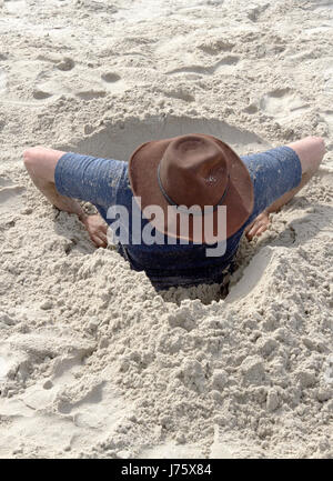 Un jeune homme tente de creuser lui-même hors d'un trou dans le sable qu'il les a en lui-même sur une plage un jour ensoleillé Banque D'Images