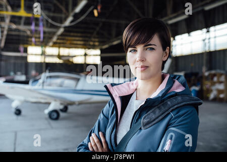 Certains jeunes femme pilote posant dans le hangar avec les bras croisés, avion sur l'arrière-plan Banque D'Images