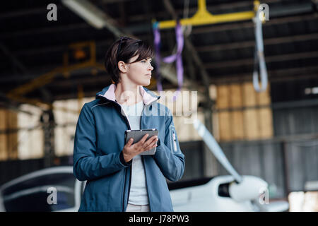 Certain femme pilote dans le hangar, elle est à l'aide d'apps sur l'aviation et à la suite de son comprimé, avion léger sur l'arrière-plan Banque D'Images