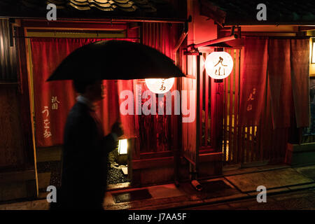 Homme tenant un parapluie passe devant une maison de thé (ochaya), dans le quartier de Gion. Les lumières donnent vie à la rue à la nuit Banque D'Images