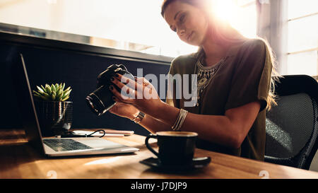 Young woman looking at camera while working on laptop. Photographe avec son appareil photo et ordinateur portable sur son bureau. Banque D'Images