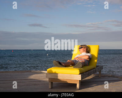 Un garçon prend une sieste sur une chaise longue jaune sur un quai pendant le coucher du soleil sur le Bitter End Yacht Club dans les îles Vierges britanniques au large des îles Banque D'Images