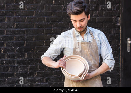 Potter, les ustensiles, la céramique art concept - male céramiste à l'avance, montre des échantillons de produits d'argile, artisan holding nouveaux bols fini, heureux maître sur fond de mur de brique gris foncé. Banque D'Images