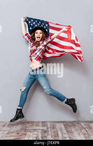 Portrait of a happy heureux jeune femme tenant un drapeau USA et sautant sur fond gris isolé Banque D'Images