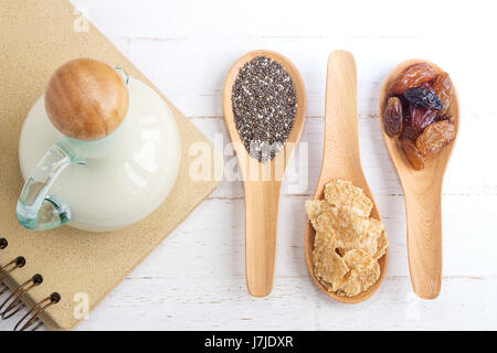 Ensemble de raisins secs, flocons de céréales de blé entier et de graines de chia dans la cuillère en bois et le lait sur fond de bois blanc Banque D'Images