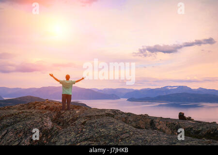 Vue panoramique sur le fjord. Le Crépuscule du temps avec le ciel rose. Le jeune homme avec les mains en l'air debout sur la falaise de roche. Banque D'Images