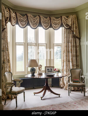 Fenêtre en baie ornées par des rideaux de soie d'Oakham par Colefax & Fowler. Une paire de meubles en bois d'un flanc fauteuils table ovale à l'avant Banque D'Images
