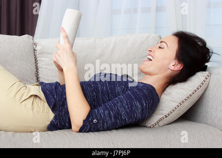 Smiling belle brune se détendre sur le canapé et la lecture d'un livre dans le salon Banque D'Images