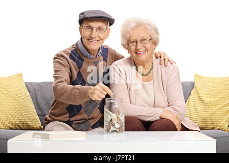 Les aînés heureux assis sur un canapé et mettre une pièce dans un pot d'argent isolé sur fond blanc Banque D'Images