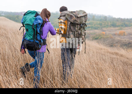 Vue arrière du jeune couple avec des sacs à dos se tenant la main et marcher dans l'herbe haute dans la campagne Banque D'Images