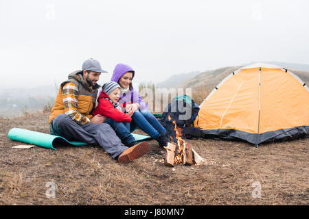 Happy Family sitting près de tente et à la recherche à un feu brûlant à nuageux jour d'automne Banque D'Images