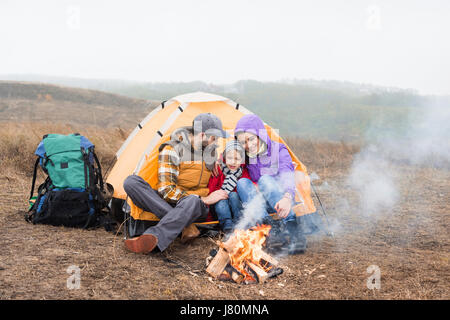 Happy Family sitting près de tente et à la recherche à un feu brûlant à nuageux jour d'automne Banque D'Images