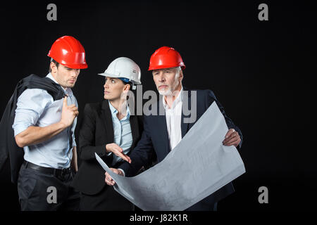 Groupe de professionnels architectes, hommes et femmes en noir sur plan discuter casques Banque D'Images