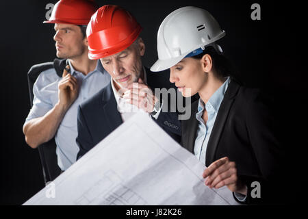 Groupe de professionnels architectes, hommes et femmes en noir sur plan discuter casques Banque D'Images