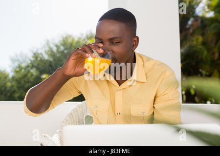 Young man drinking orange juice while looking at laptop in cafe Banque D'Images