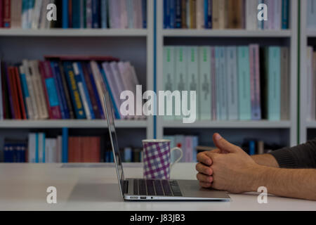 Guy ou l'homme d'affaires ou étudiant ou travailler sur un ordinateur portable dans une bibliothèque. Étagères en arrière-plan et tasse de café à côté de lui Banque D'Images