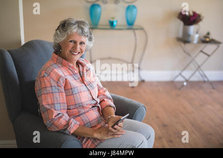 Portrait of smiling senior woman holding mobile phone, assis sur un fauteuil à la maison Banque D'Images