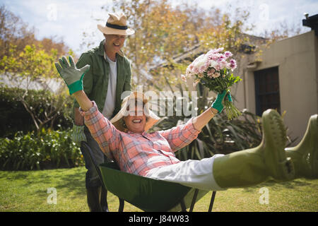 Man carrying cheerful senior woman holding bouquet à emprunter à roue arrière-cour Banque D'Images
