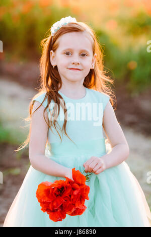 Kids Fashion, enfance heureuse, mariage, nature concept - portrait de jeune femme charmante en robe turquoise avec des coquelicots rouges dans sa main et s'écoulant les cheveux bruns, elle est debout dans la lumière du soleil Banque D'Images