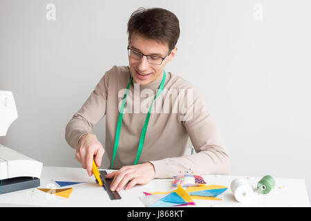 Quilting et patchwork à l'atelier - jeune homme assis au bureau sur mesure et de découper des pièces de tissus pour patchwork couture articles, allongé près de bobines de fil et morceaux de tissus. Banque D'Images