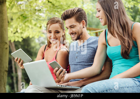 Les élèves heureux dans le parc de détente sur un banc et à l'aide d'un ordinateur portable, une tablette numérique et un téléphone intelligent Banque D'Images