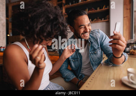 Smiling young couple sitting at coffee shop à l'aide de téléphone mobile. Jeune homme et femme à prendre café et selfies rire. Banque D'Images