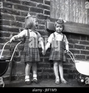 Années 1950, historiques, deux petites filles, sœurs, portant des robes tartan correspondant avec leur poupée jouet poussettes à l'extérieur d'une maison en brique, un rire, se tenant la main. Banque D'Images