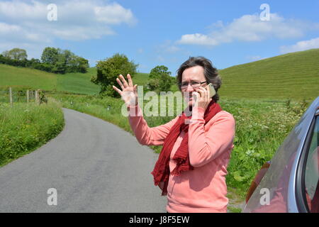 Femme debout par la papeterie voiture sur un chemin de campagne, à l'aide de téléphone mobile pour demander de l'aide Banque D'Images