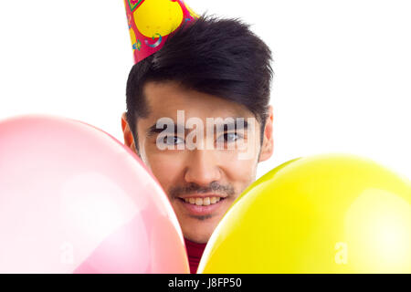 Jeune homme souriant aux cheveux noirs en chemise rouge avec célébrer hat holding beaucoup de ballons colorés sur fond blanc en studio Banque D'Images