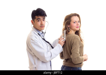 Beau jeune médecin aux cheveux noirs en robe blanche avec stéthoscope à l'écoute de l'arrière de jeune femme agréable avec de longs cheveux bruns en chandail et Banque D'Images