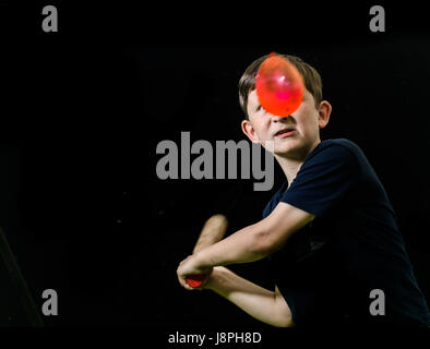 Jeune garçon, avec un regard de détermination et la concentration sur son visage, s'attaque avec son bat à un ballon d'eau rouge avec l'intention de craquer il Banque D'Images