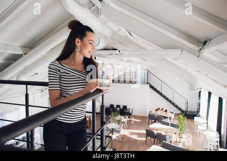 Portrait de femme debout avec une tasse et en regardant les gens de l'autre étage. Femme adulte heureux à l'écart de l'appareil photo. Banque D'Images