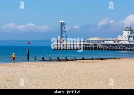 Sauveteur RNLI femme marchant le long de la mer à la plage de Bournemouth avec jetée dans la distance en mai Banque D'Images