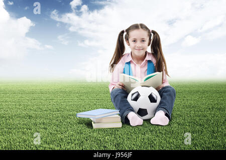 Cute little girl avec des livres et d'un ballon de football sur l'herbe verte Banque D'Images