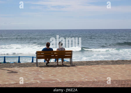 Couple en vacances s'asseoir et se détendre sur un banc de bois qu'ils en ont l'air de l'océan Atlantique à partir de la promenade de Playa Las Americas à Teneriffe dans th Banque D'Images