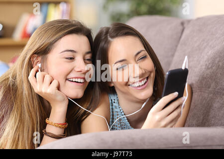 Vue avant portrait de deux amis joyeux à l'écoute de la musique en ligne à un téléphone intelligent allongé sur un canapé dans la salle de séjour à la maison Banque D'Images