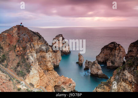 Photographe sur haut des falaises entouré par la mer sous le ciel rose au lever du soleil, Ponta da Piedade, Lagos, Algarve, Portugal Banque D'Images