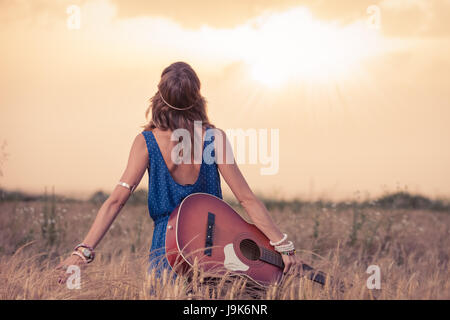 Jeune femme de style hippie rétro avec guitare acoustique en champ de blé à sun à trouver de l'inspiration pour la chanson suivante. La musique, l'art et le style de conc Banque D'Images