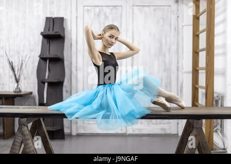 Ballerine Attrective s'assied sur la table en bois sur le mur de lumière dans le studio. Elle porte un justaucorps noir cyan avec un tutu et pointes. Banque D'Images