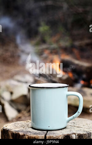 L'émail bleu tasse de café fumant assis sur un vieux journal par un feu de camp en plein air. L'extrême profondeur de champ avec selective focus on chope. Banque D'Images