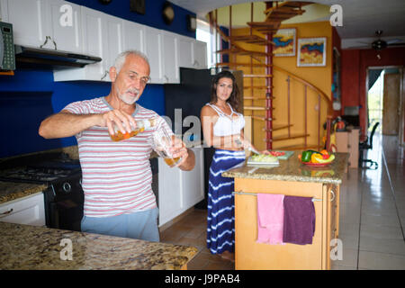 Homme plus âgé se verse un verre de bière pendant que la jeune femme prépare le dîner dans la cuisine Banque D'Images