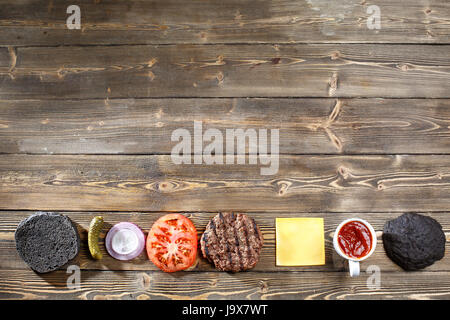 Ingrédients pour la cuisson burger noir. La viande grillée patty, brioches, oignons, tomates, laitue, cornichons, sauce, fromage sur fond de bois noir Vue de dessus Banque D'Images