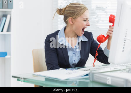 Jeune femme crier en direction du récepteur téléphonique rouge at desk in office Banque D'Images