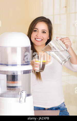 Belle femme tenant un verre d'eau dans une main et un pichet d'eau dans son autre main, avec un système de filtre de purificateur d'eau sur un arrière-plan de cuisine Banque D'Images