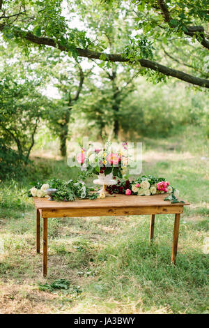 Maison de vacances d'été, de fleurs et d'arrangement de fleurs concept - magnifique bouquet dans vase blanc sur la table en bois, blanc et jaune rose roses, pivoines, œillets rouges, les fleurs coupées, glade parmi les arbres. Banque D'Images