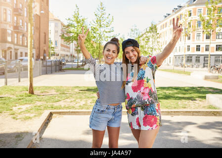 Deux jeunes femmes donnant confiance mignon V-signes tels qu'ils sont main dans la main souriant à la caméra dans un milieu urbain sur une journée ensoleillée Banque D'Images