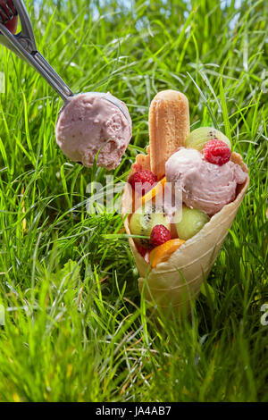 Fruité de la crème glacée dans un cornet gaufré avec des biscuits, des framboises et des tranches de fruits à proximité d'une cuillère en métal avec de la glace. Une composition sur fond de g Banque D'Images