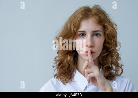 Une rousse, jeune fille aux cheveux bouclés avec rousseur met le doigt sur ses lèvres. Un signe de silence ou secret. Banque D'Images