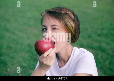 Très beau portrait model eating pomme rouge dans le parc. Portrait de l'extérieur, jolie jeune fille assise sur l'herbe verte Banque D'Images