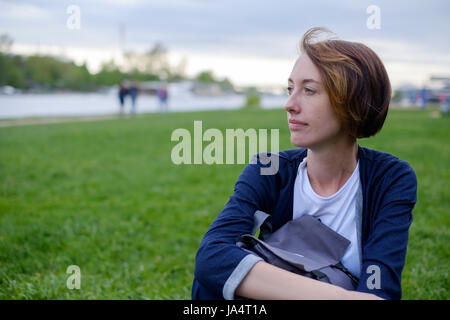 Une fille avec des taches de rousseur est assis sur l'herbe verte dans le parc. Elle est titulaire d'un sac à dos dans ses mains. se reposer à l'été dans l'air frais. Banque D'Images